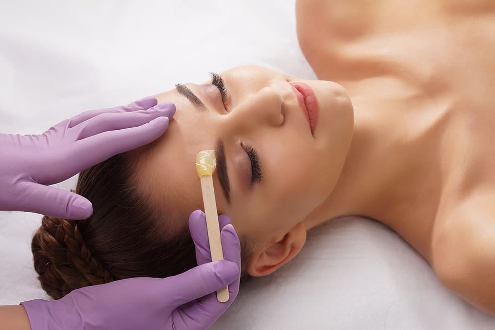 Beauty therapist applying warm wax to a client's eyebrow area with a wooden spatula during an eyebrow re-shape and wax treatment at The Zen Den.