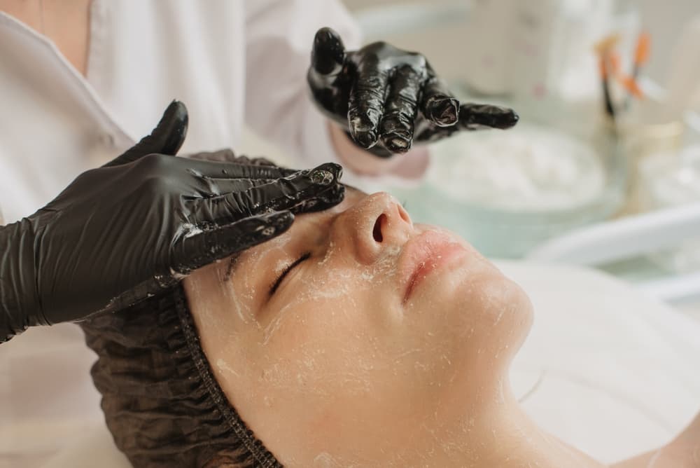 Beauty therapist wearing black gloves applying a fruit peel facial treatment to a client's face at The Zen Den, focusing on exfoliation and skin renewal.