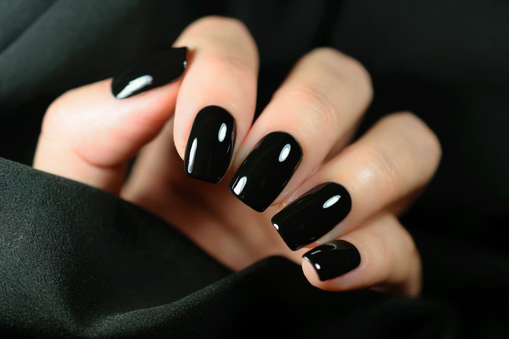 Close-up of a hand with sleek, glossy black gel polish, nails shaped square and evenly finished, displayed against a soft black fabric background, representing The Zen Den's gel polish hands service.