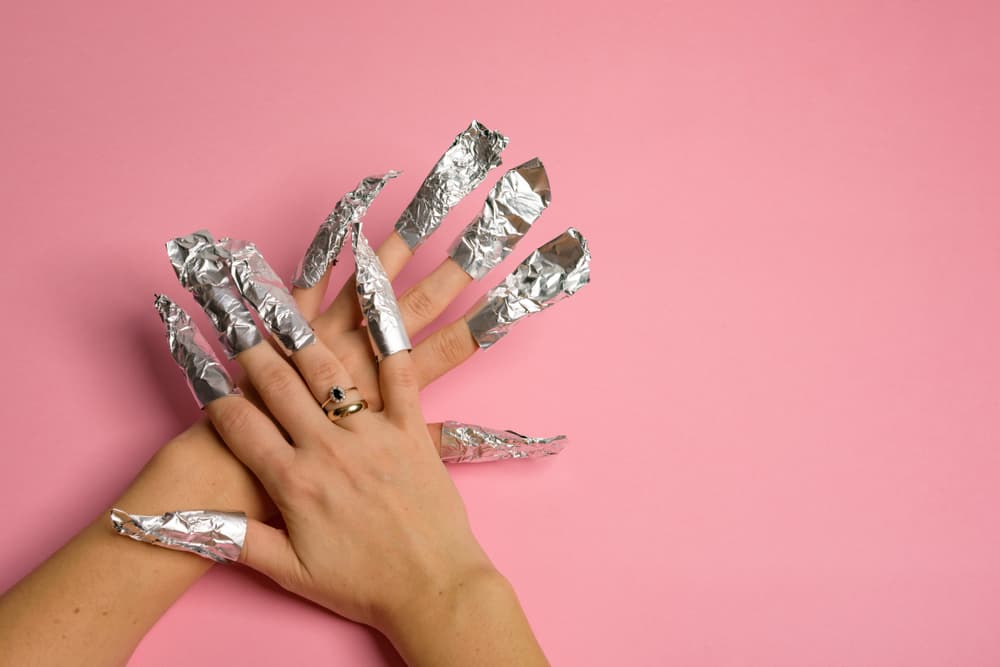 Hands wrapped in aluminium foil nail wraps during the gel polish removal process, set against a pink background.