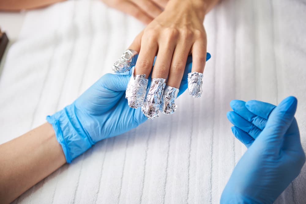 Professional gel polish removal at The Zen Den, showing hands with foil wraps on nails applied by a therapist wearing blue gloves.
