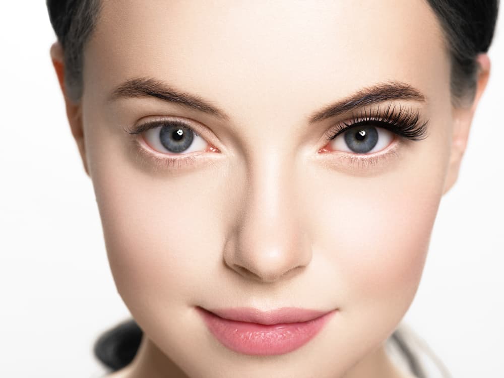 Close-up of a woman's face showing a comparison of natural brows and lashes on one side and enhanced, fuller hybrid brows and lashes on the other, highlighting the dramatic difference.