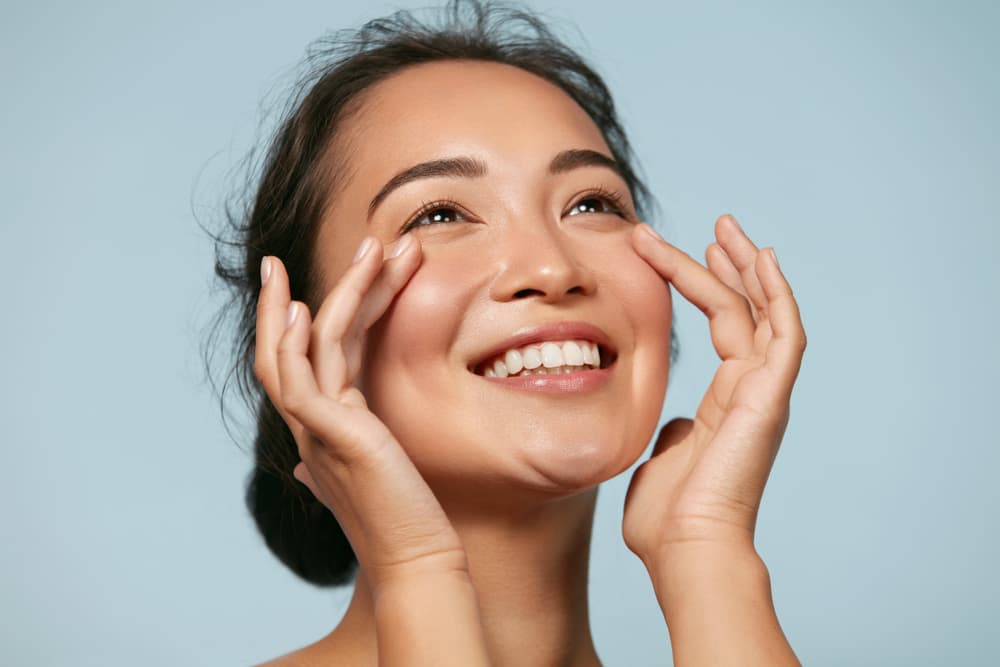 Smiling woman with radiant, hydrated skin gently touching her face after a Hydrate Facial treatment at The Zen Den.