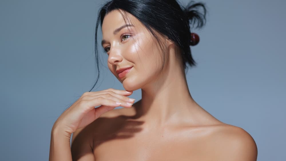 Beautiful woman with glowing, healthy skin posing against a soft grey background, showing the results of The Zen Den’s In-House Classic facial treatment for refreshed and radiant skin.