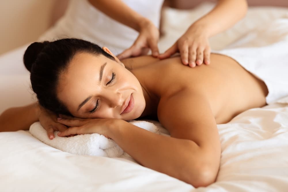 A peaceful woman lying on a massage table with her head resting on a towel, eyes closed, as a therapist performs a gentle and relaxing back massage at The Zen Den.