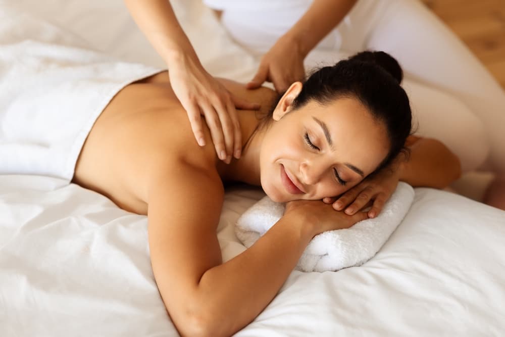 A relaxed woman lying face down on a massage table with a towel under her head, smiling as a therapist performs a soothing neck and shoulder massage at The Zen Den.