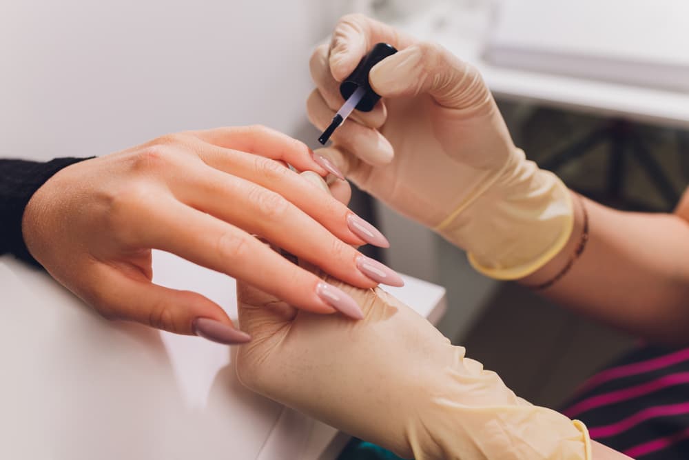 Close-up of a beauty therapist applying nude pink gel polish to well-shaped nails during a luxury gel manicure, with gloves worn for hygiene and precision.