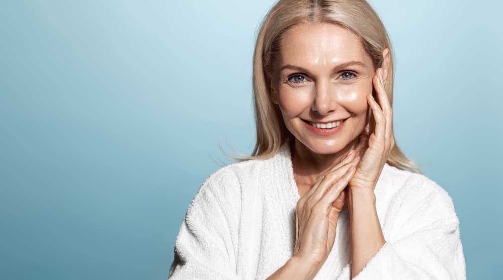 Smiling mature woman with healthy, radiant skin touching her face after a Nourish Facial treatment at The Zen Den.