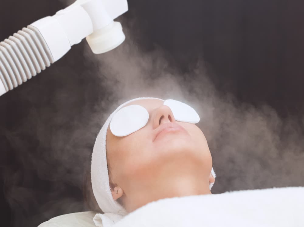 Woman relaxing with eye pads during an ozone steam facial treatment at The Zen Den, with warm steam directed onto her face for deep cleansing and hydration.