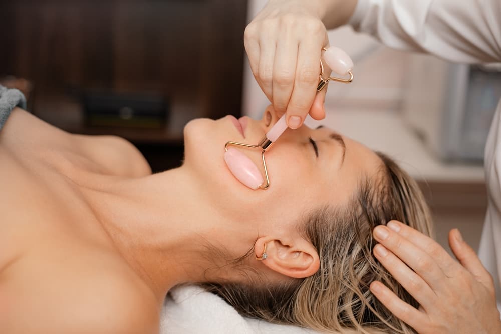 Woman receiving a relaxing Rose Quartz Crystal Facial at The Zen Den, with a therapist using a pink quartz roller to massage the jawline and promote skin radiance.