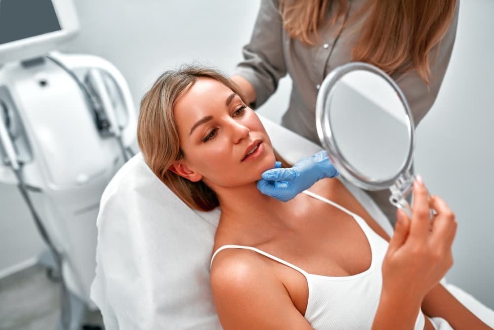 Woman sitting in a treatment chair holding a mirror while a skincare specialist wearing gloves examines her face during a Tropic Skincare consultation at The Zen Den.