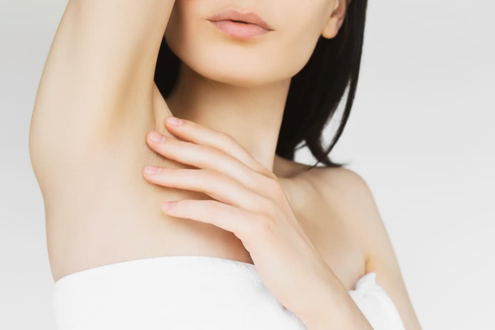 Close-up of a woman with smooth skin raising her arm to reveal a hair-free underarm, wearing a white towel wrap, symbolising the results of professional underarm waxing.