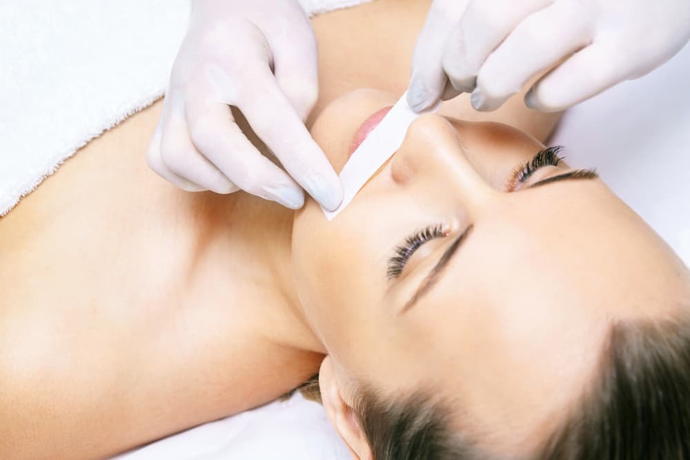 Woman lying down during a professional beauty treatment as a therapist wearing gloves applies a waxing strip to the upper lip area for hair removal.