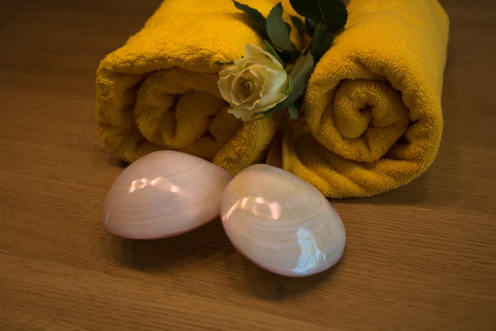 Two rolled yellow towels with a cream rose and smooth polished shells arranged on a wooden surface, representing the warm massage for two experience at The Zen Den.