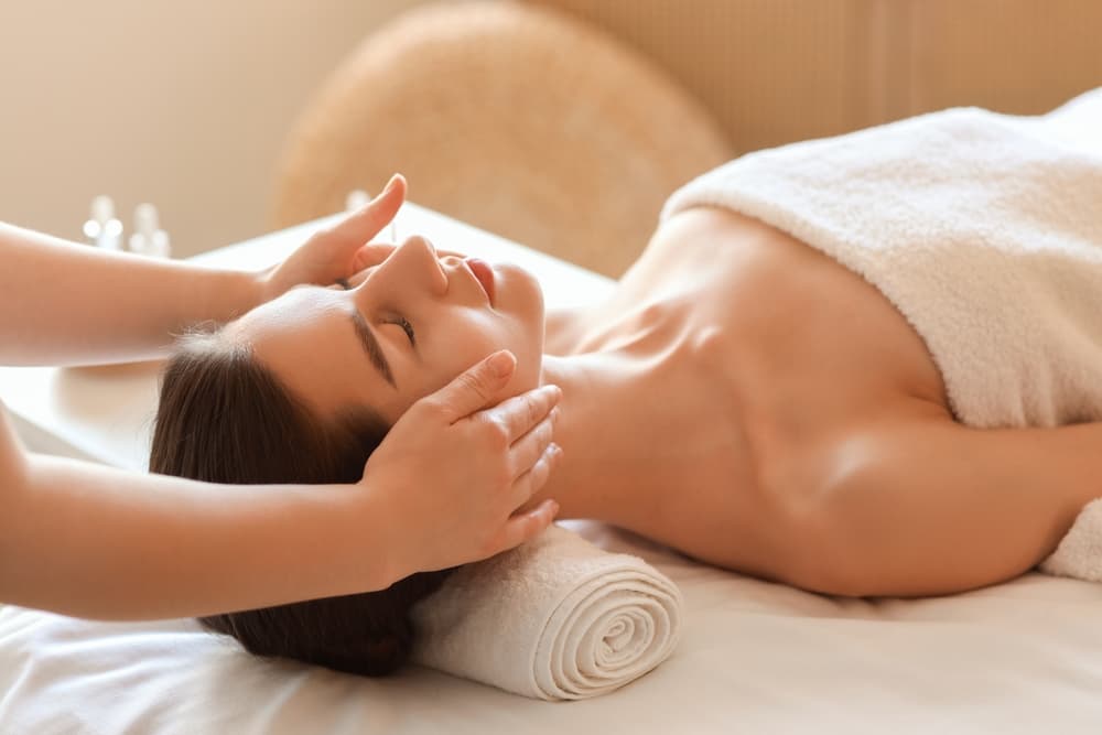Relaxed woman lying on a massage table with a towel covering her, receiving a gentle face and head massage in a serene spa setting.